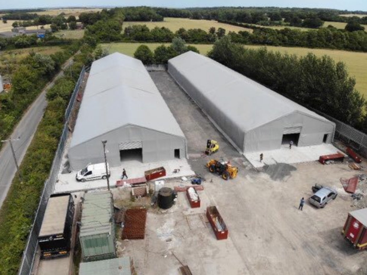 Aerial view of two large, gray industrial fabric buildings in a construction yard with vehicles and equipment.