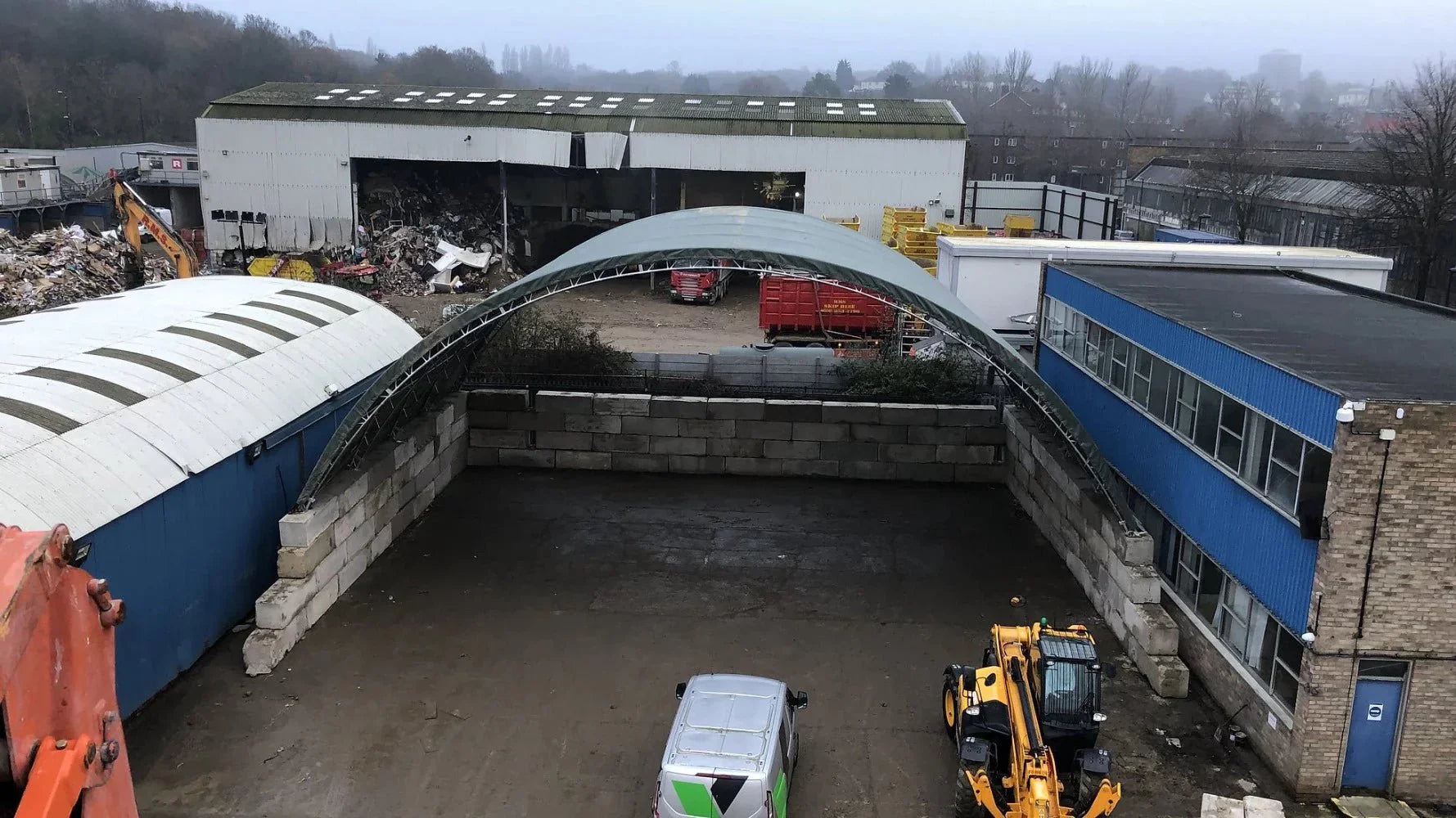 Industrial area with machinery, storage buildings, and recycling materials under an overcast sky.