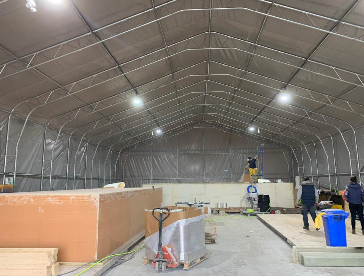Large building interior with a metal frame, two people, a large wood object, and one person on a tall ladder.