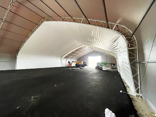 Interior of a large, white, arched fabric structure. Black flooring, metal frame. Construction equipment visible in the distance.
